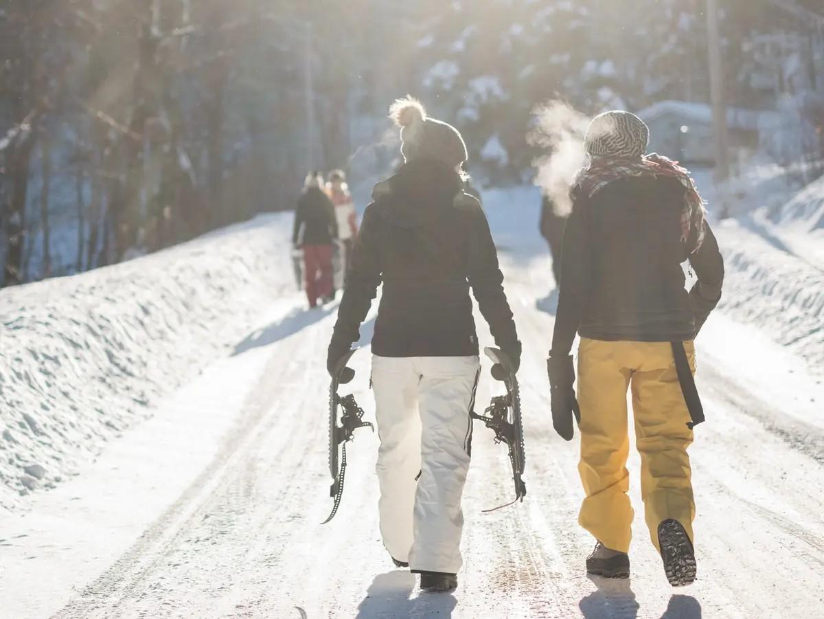 Zwei Menschen mit Skiern gehen nebeneinander eine geräumte Straße entlang