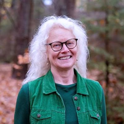 Iris Bein, eine Frau mit weißen Haaren, Brille und grünem Outfit lächelt in die Kamera, im Hintergrund herbstlicher Wald.