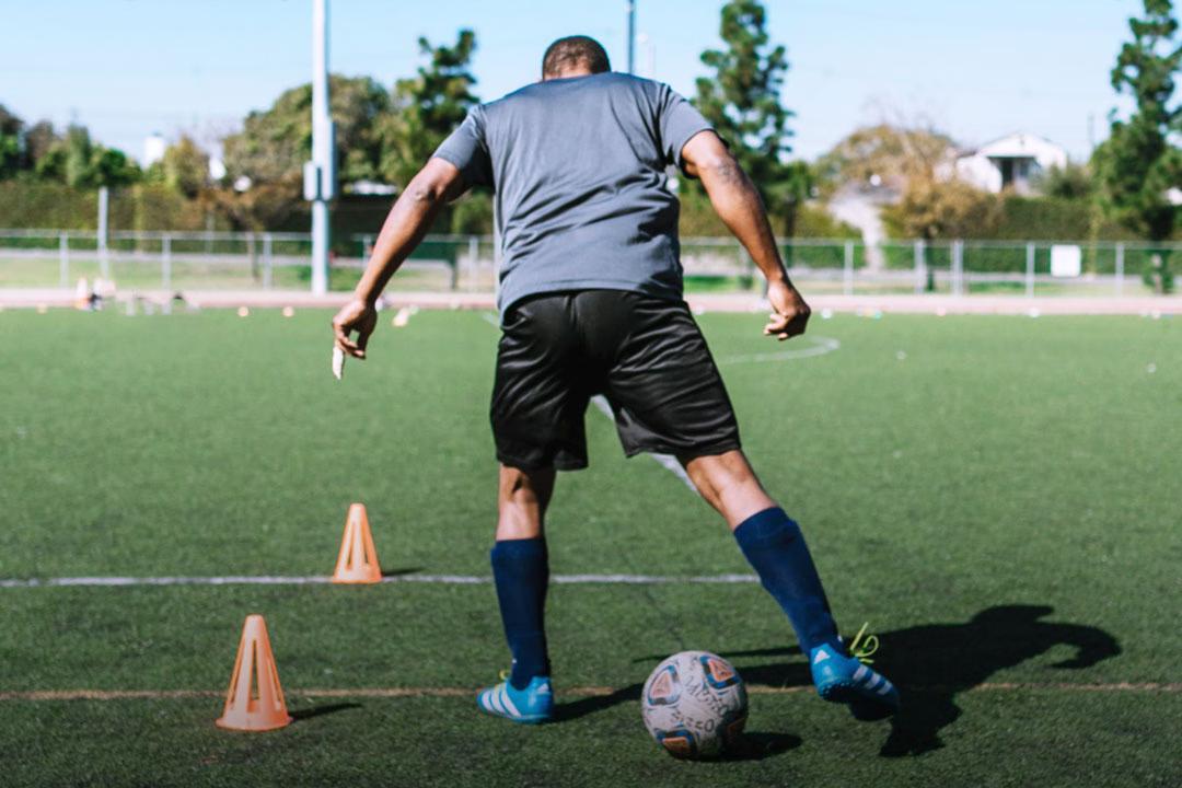 Ein Fußballtrainer läuft auf Rasen mit einem Fußball um orangefarbene Hütchen herum, den Rücken zur Kamera gewandt.