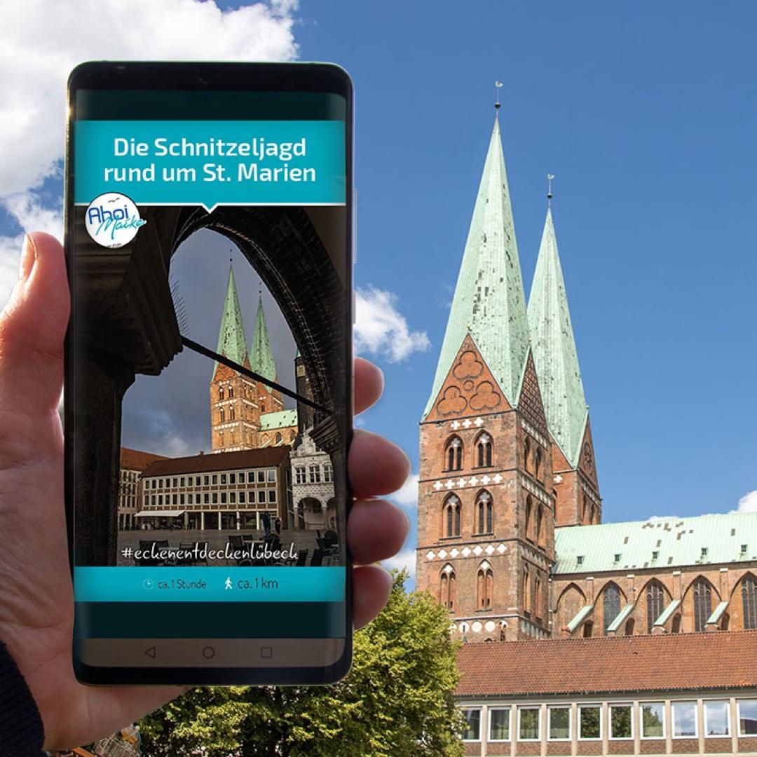 Smartphone zeigt Schnitzeljagd rund um St. Marien in Lübeck, im Hintergrund die Kirche mit grünem Doppelturm.
