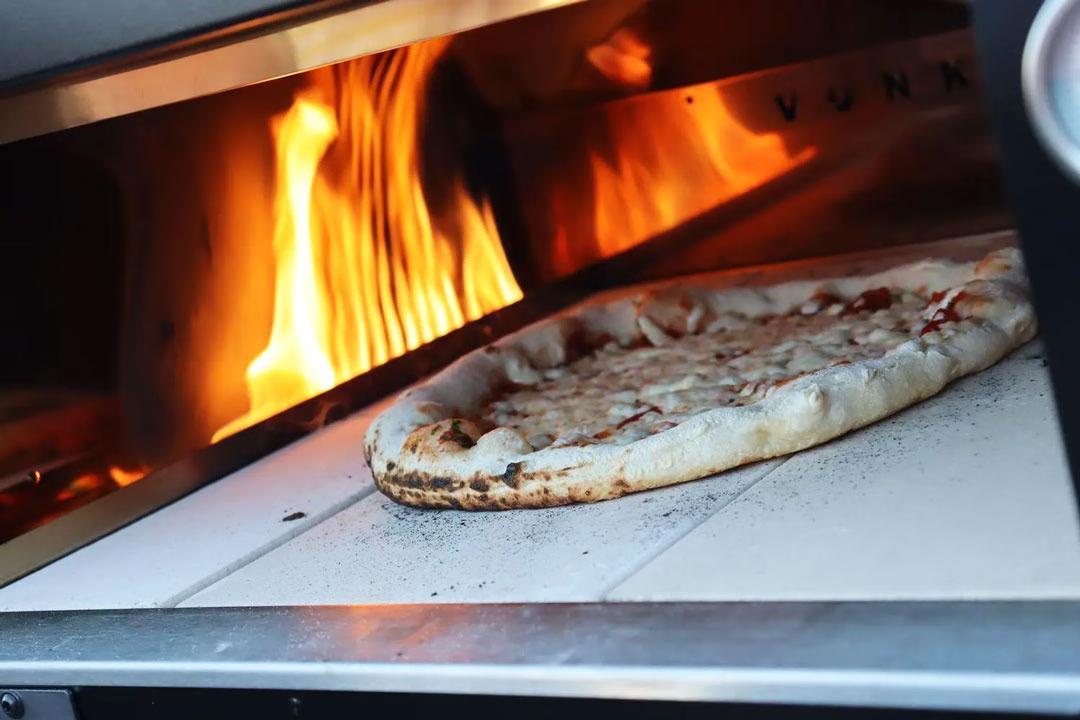 Pizza backen im Steinofen: Frisch gebackene Pizza mit perfektem Rand vor loderndem Feuer im Pizzaofen.