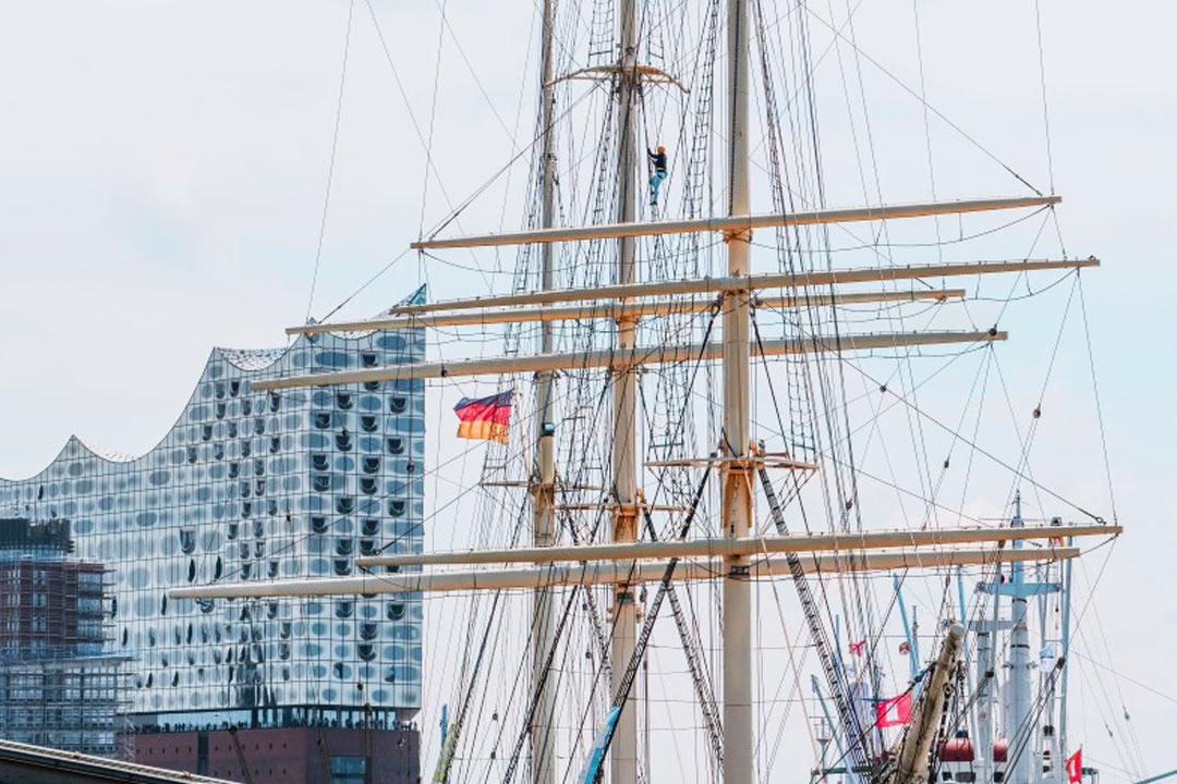 Kletterer im Mast der Rickmer Rickmers mit Elbphilharmonie im Hintergrund im Hamburger Hafen.