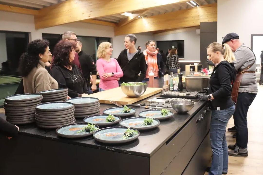 Mehrere Menschen stehen in der Eventküche beim Afterwork Burger-Grillkurs und schauen dem Profikoch zu.