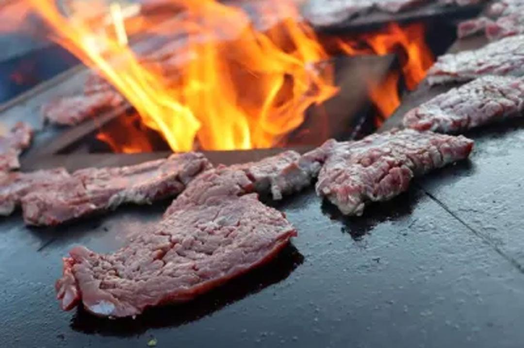 Saftige Steakscheiben auf heißem Grillstein mit lodernden Flammen im Hintergrund.