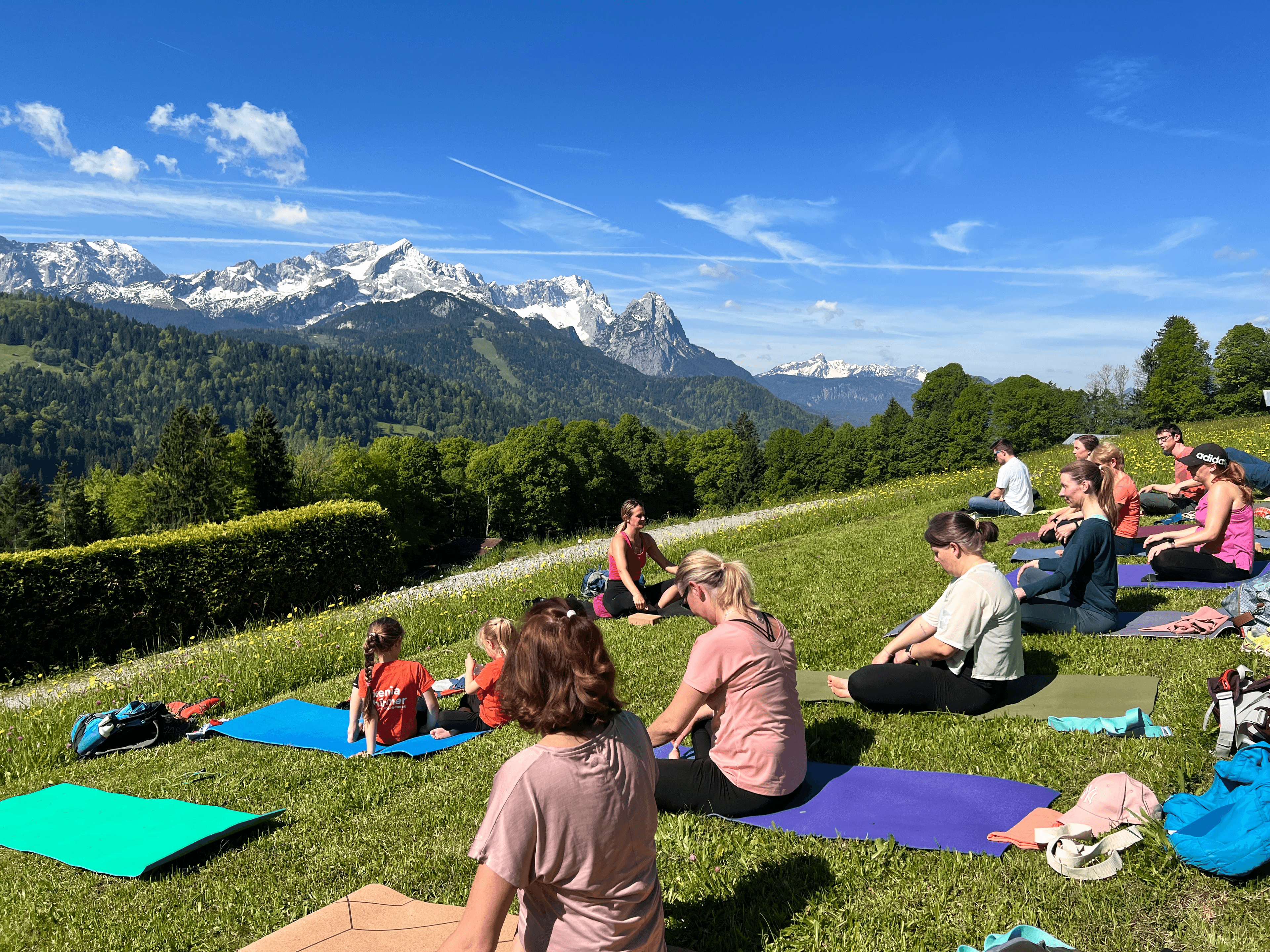 Teilnehmende sitzen beim Yoga in den Bergen im Schneidersitz, im Hintergrund sind die Alpen zu sehen