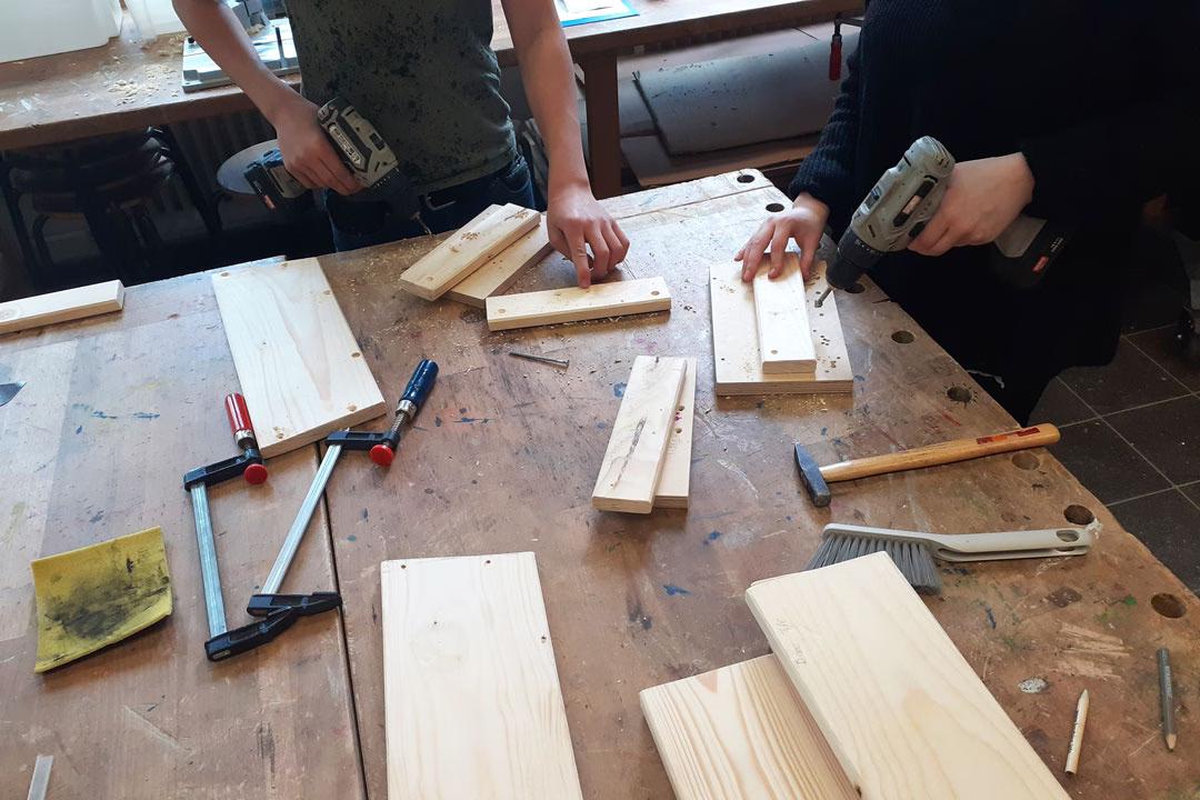 Zwei Personen bauen in der Holzwerkstatt an einem Regal und arbeiten mit Akkuschrauber, Hammer und Schraubzwingen.