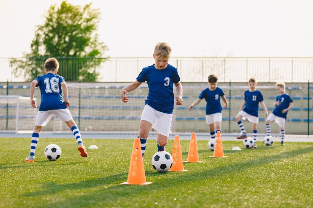 Mehrere Jungs spielen Fußball und dribbeln Fußbälle um orangefarbene Pylonen auf einem Rasenplatz.