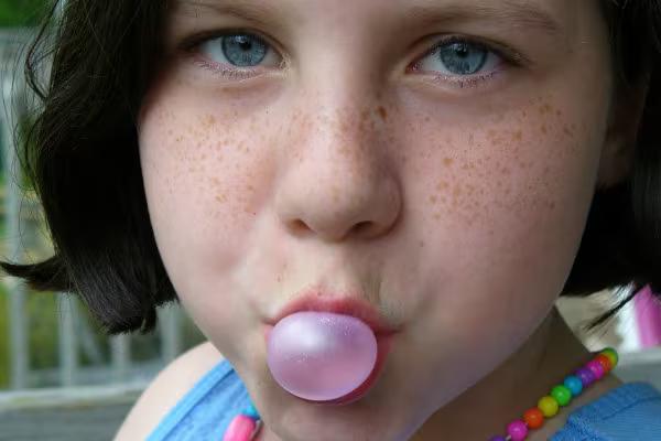 Nahaunahme eines Mädchens mit dunklen Haaren, das ein rosa Kaugummi aufbläst.