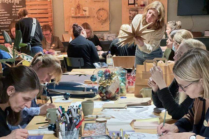 Viele Personen sitzen an mehreren Tisch in der Machbar in Lübeck und basteln kreativ mit Stiften, Papier und Rohlingen.