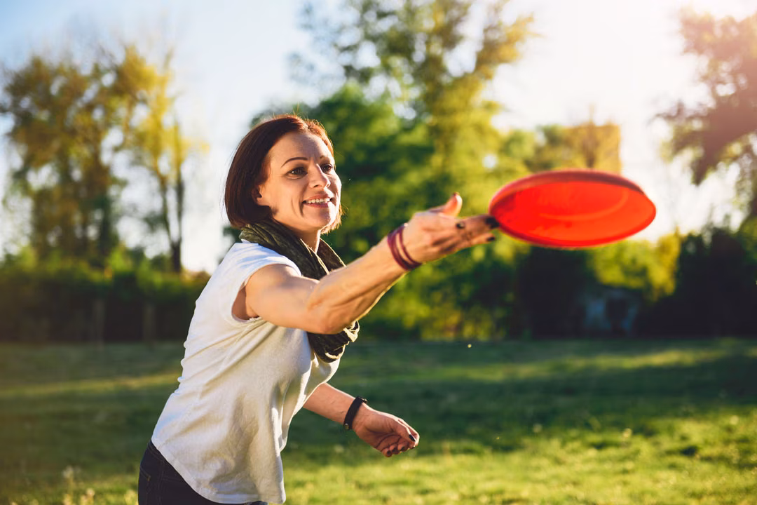 Lächelnde Frau spielt Frisbee Golf und wirft eine rote Scheibe auf einer grünen Wiese im Sonnenschein.