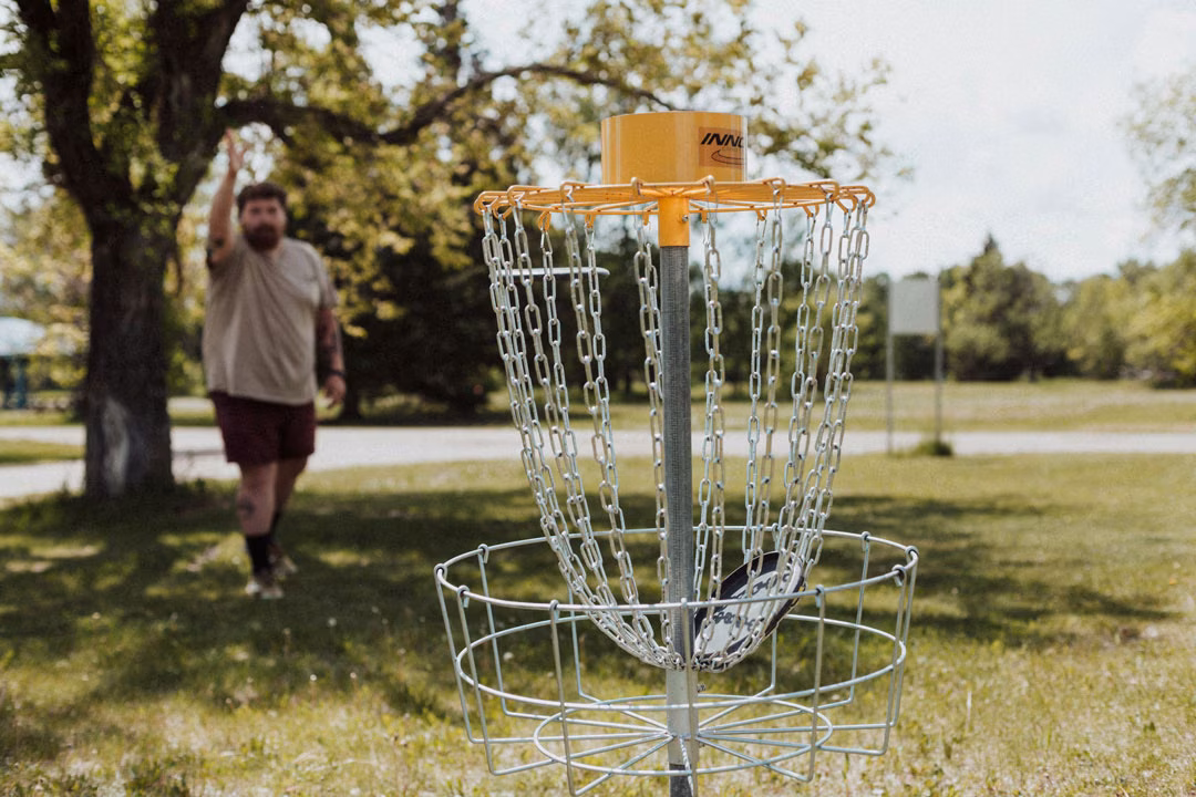 Mann mit Bart spielt Disc Golf und wirft eine Scheibe Richtung Korb im Park.