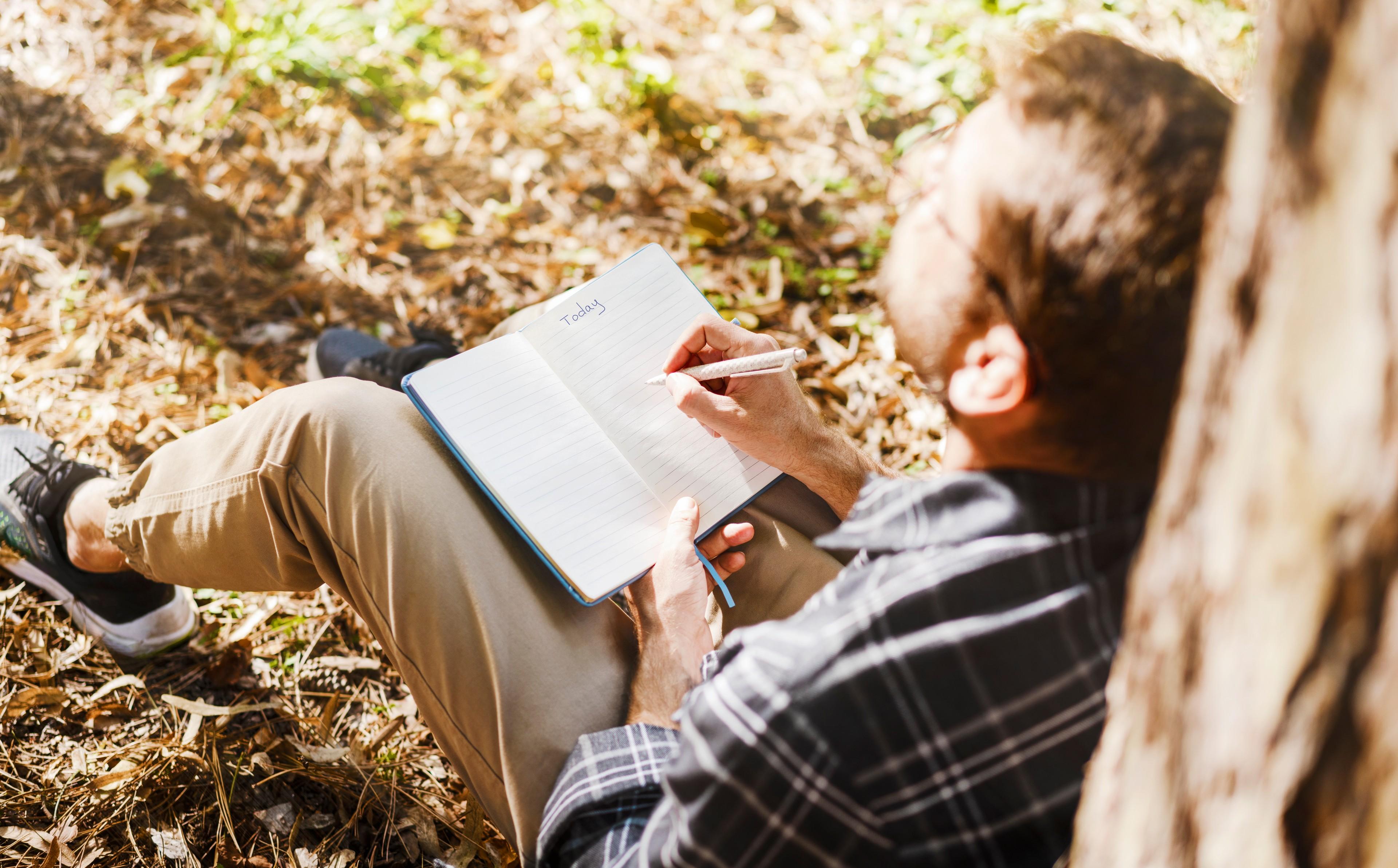 Ein Mann sitzt bei einem Wald-Retreat an einen Baumstamm gelehnt und schreibt in ein Journal