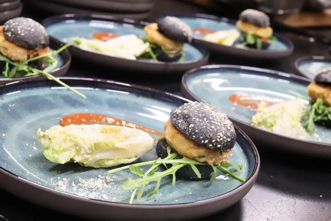 Mehrere Teller mit schwarzen Burger-Buns mit Fleisch-Patties, dazu ein gebratener Kohlkopf und Rucola, daneben Sauce.