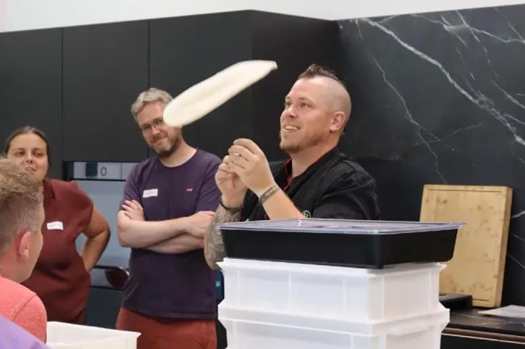 Pizza backen lernen: Pizzabäcker demonstriert Teigwerfen im Pizzakurs mit begeisterten Teilnehmer:innen.