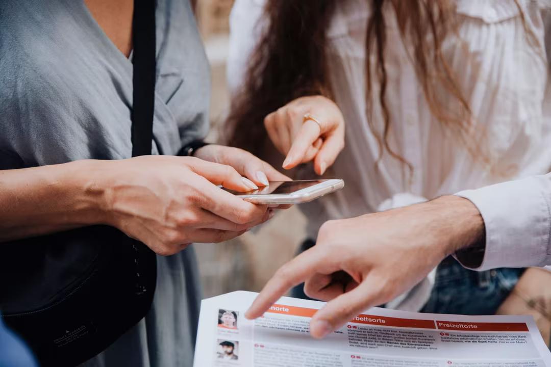 Zwei Personen schauen in ein Smartphone, eine weitere Person zeigt auf die ausgedruckten Unterlagen des Krimi-Trail Grömitz.