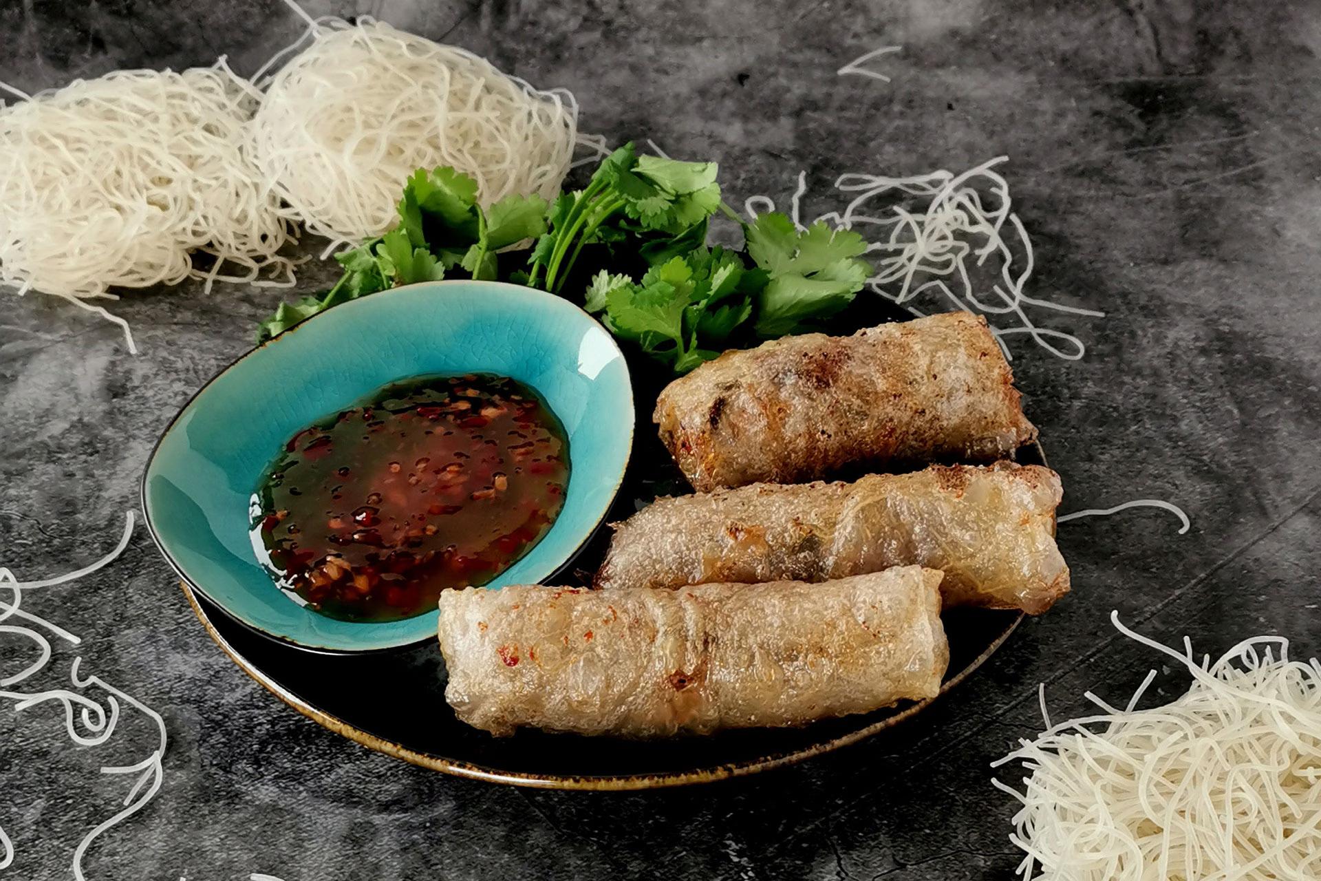 Authentisches Gericht aus Vietnam: Frühlingsrollen mit Ponzu-Sauce auf einem Teller, daneben Koriander und Reisnudeln.