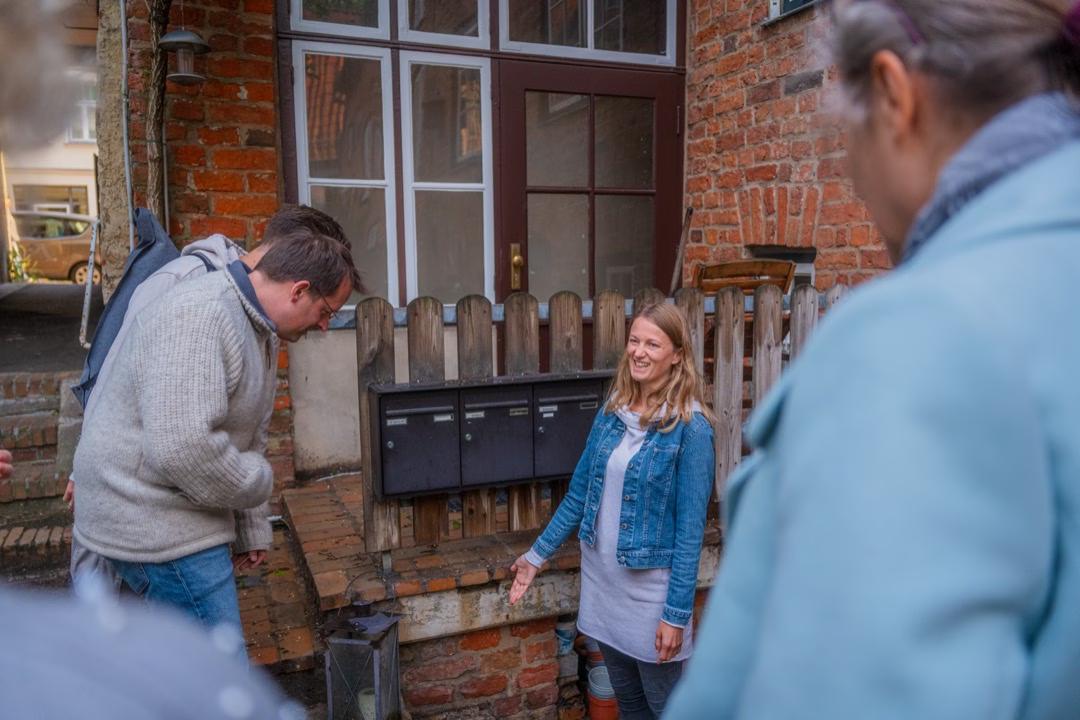 Eine Frau steht vor einer Briefkastenreihe an einem Backsteingebäude in Lübeck & erklärt etwas lächelnd ihren Zuhörer:innen.