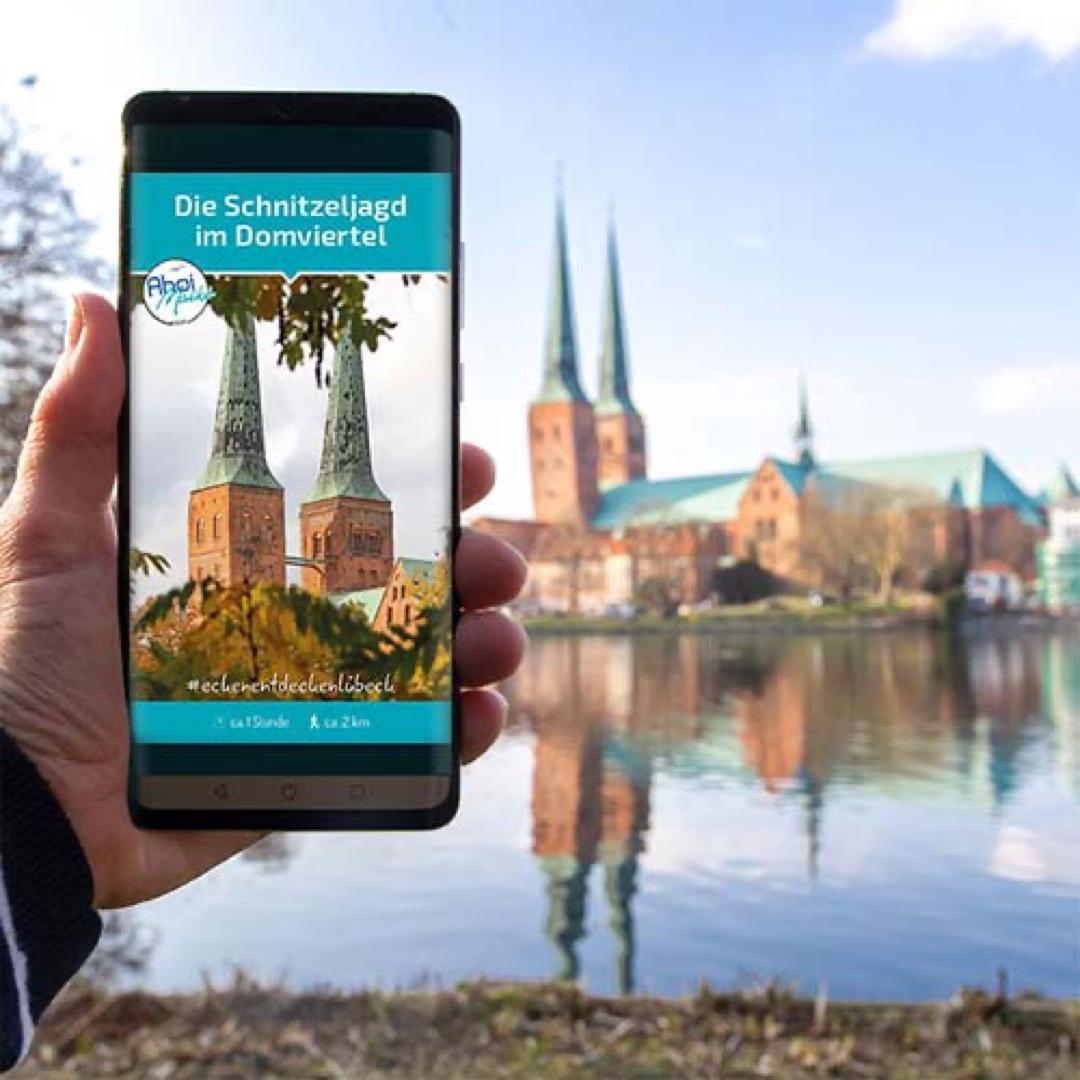 Smartphone zeigt Schnitzeljagd im Domviertel in Lübeck, im Hintergrund spiegeln sich die Domtürme im Wasser.