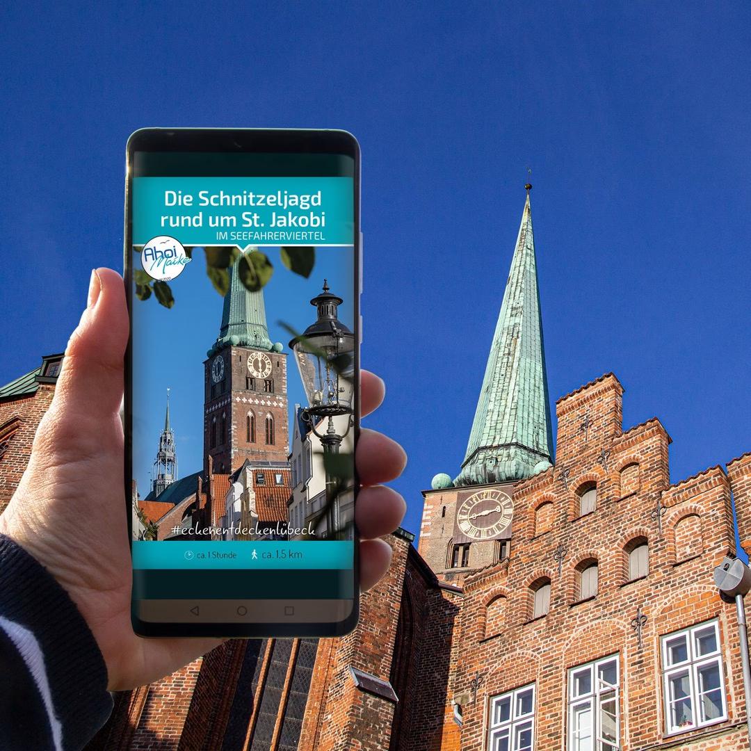 Smartphone zeigt Schnitzeljagd rund um St. Jakobi in Lübeck, im Hintergrund die Kirche mit grünem Turm.