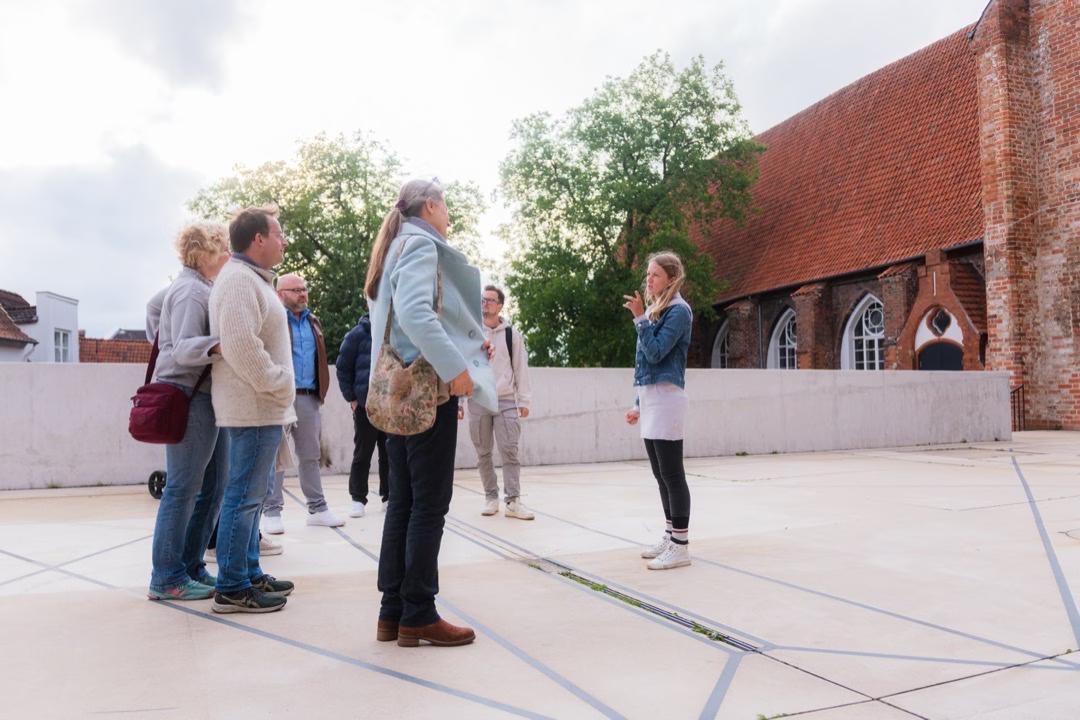 Eine Stadtführerin steht auf einem hellen Kirchenvorplatz und redet mit der vor ihr stehenden Gruppe.