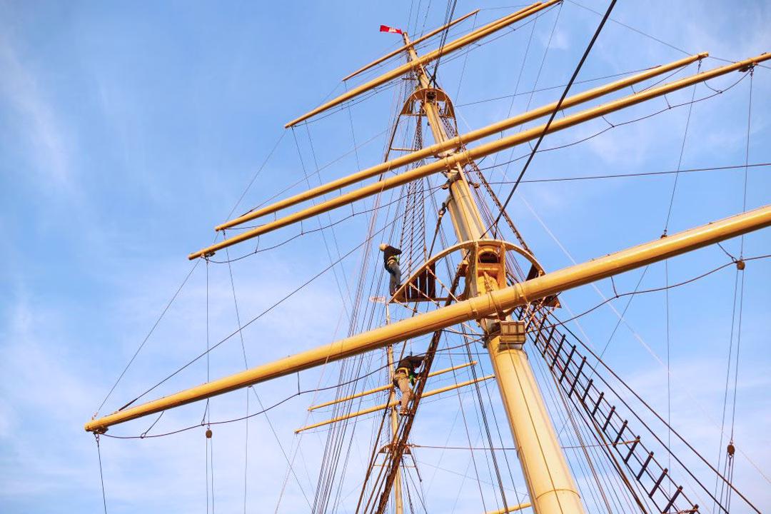Zwei Personen steigen gesichert im Mast der RICKMER RICKMERS bei blauem Himmel empor.
