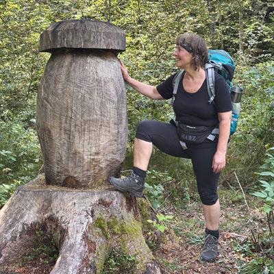 Frau mit Rucksack wandert im Wald und posiert lachend neben einer großen hölzernen Skulptur in Form eines Pilzes.
