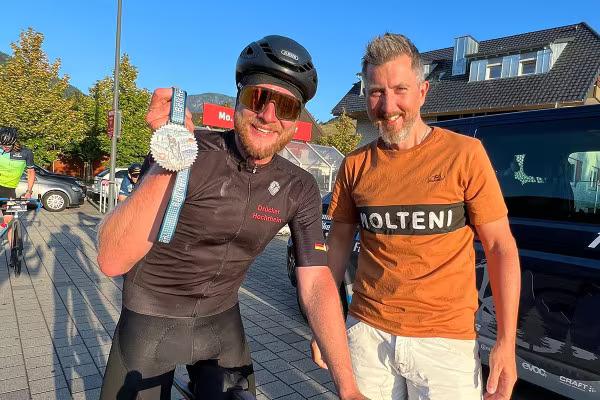 Ein Mann in Rennradkleidung auf seinem Rennrad hält seine Medaille beim Belchen Hoch 3 in die Kamera. Daneben ein Freund.