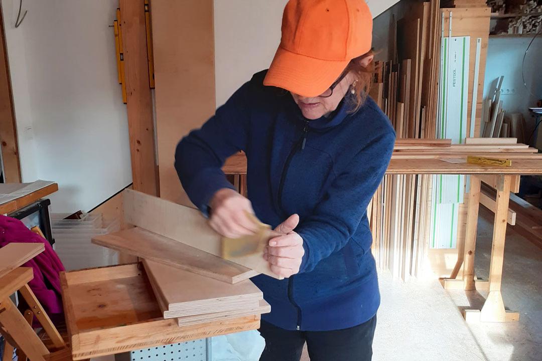 Frau beim Mädelsabend in der Holzwerkstatt: Sie trägt Kappe und schleift konzentriert ein Holzbrett für ihr eigenes Unikat.