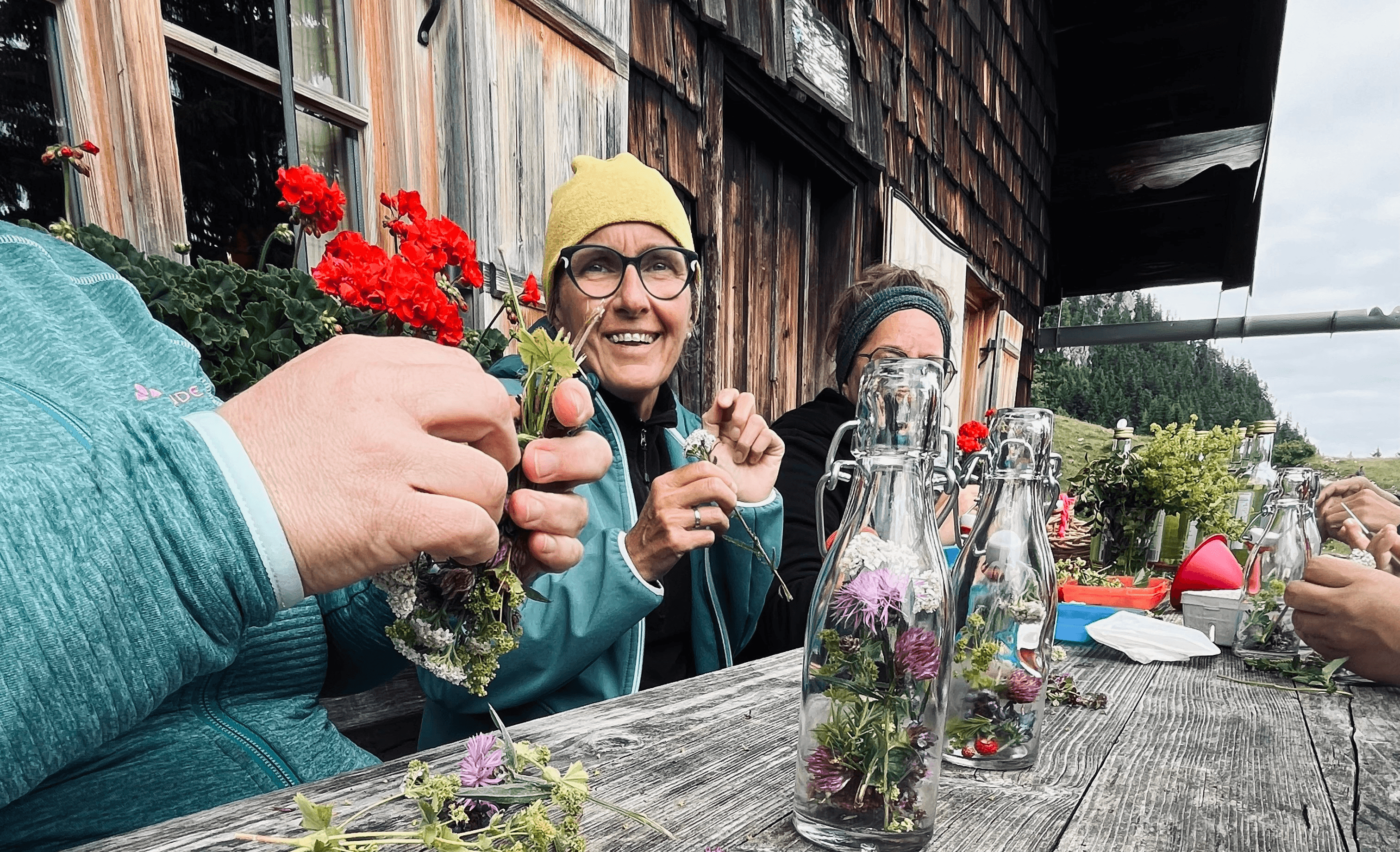 Teilnehmende eines Kräuterkochkurses sitzen an einem Holztisch vor einer Alm und verarbeiten Wildkräuter