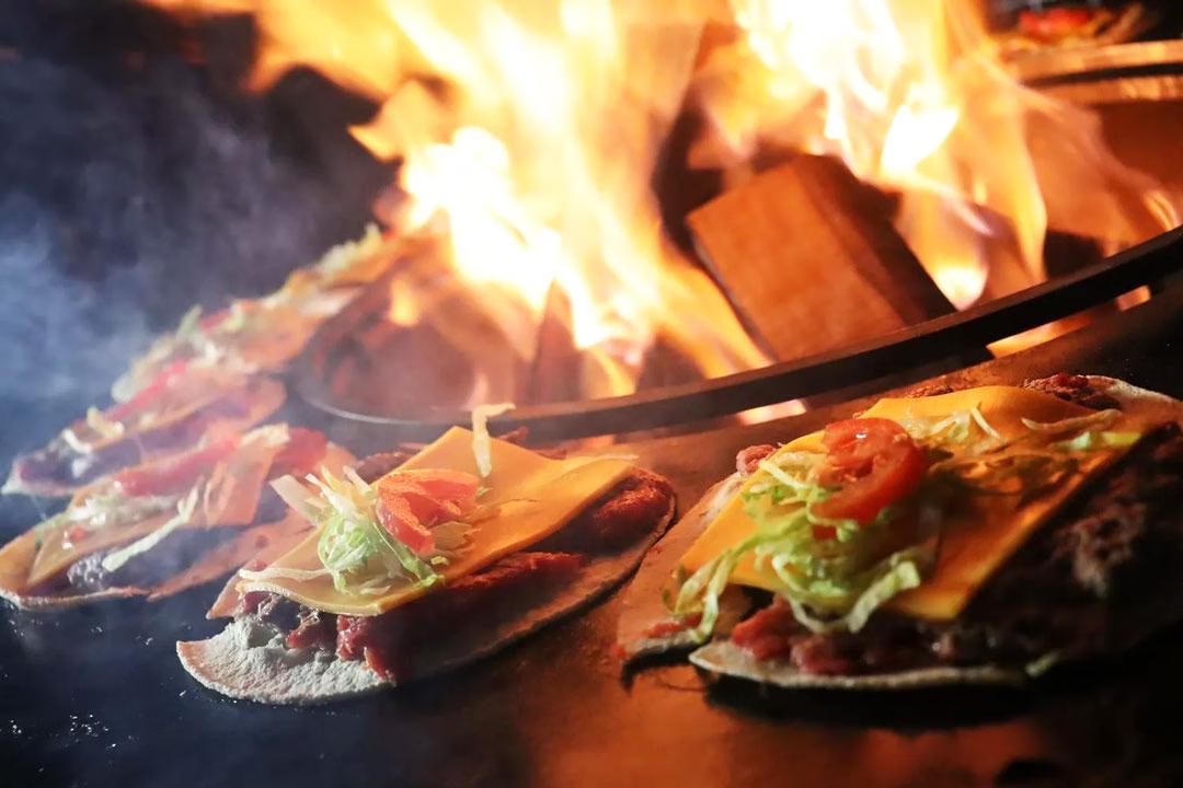Mehrere Tacos auf gusseisernem Feuerring beim Afterwork Burger-Grillkurs. Im Hintergrund lodert das Feuer.
