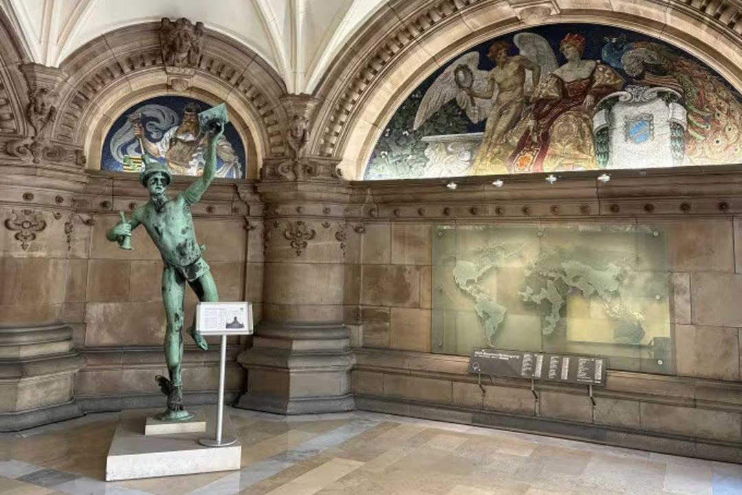 Statue in der historischen Bremer Baumwollbörse, flankiert von bunten Mosaiken und einer Weltkarte an der Wand.