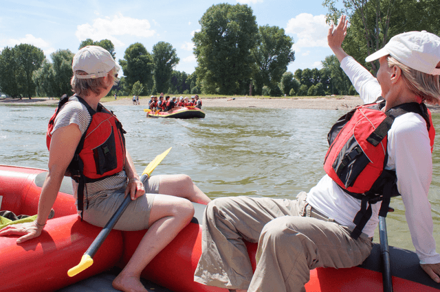 Teilnehmende einer Rafting-Tour winken einem anderen Boot auf dem Rhein in Köln