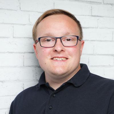 Ein Porträtfoto von Marcel Bodien, Fußballtrainer mit schwarzer Brille und schwarzem Polo-T-shirt vor einer weißen Wand.