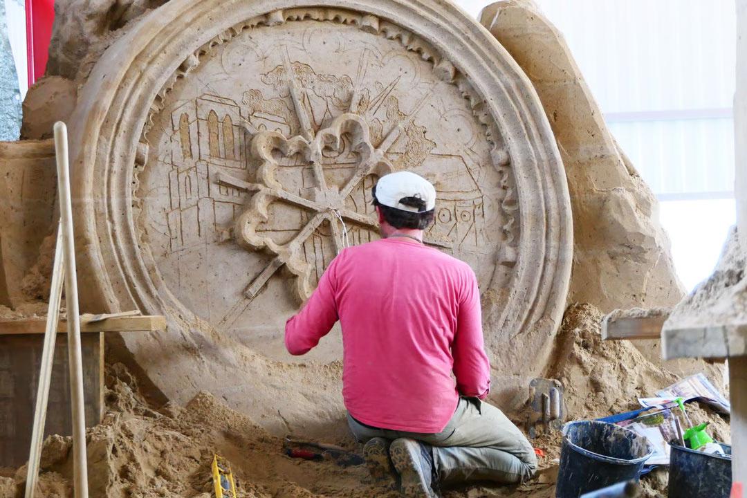 Sandskulpturenausstellung: Künstler kniet und bearbeitet ein rundes Reliefmedaillon mit Stadtmotiven aus Sand.