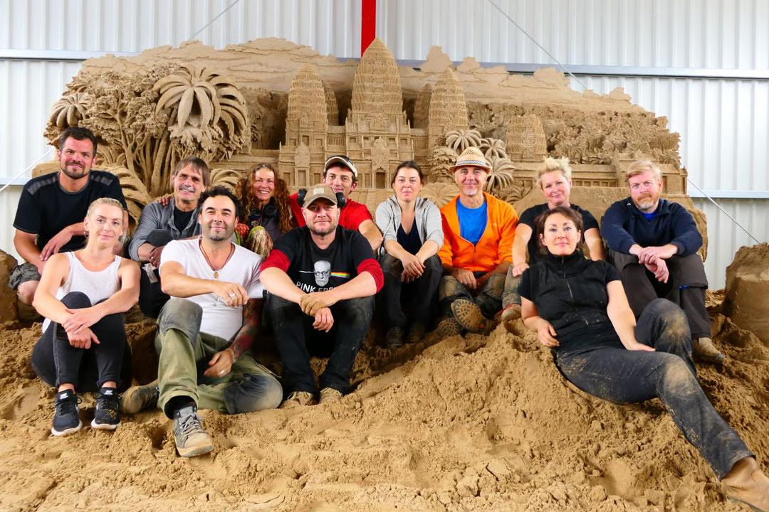 Sandskulpturenausstellung: Zwölf Künstler:innen posieren vor einer Sandskulptur des Angkor Wat Tempels.