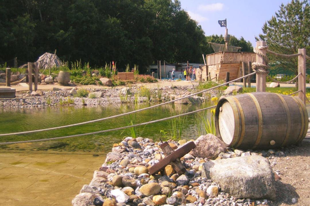 Ein Fass vor einem Teich, im Hintergrund das Piratenschiff der Minigolf-Anlage Piraten der Ostsee.