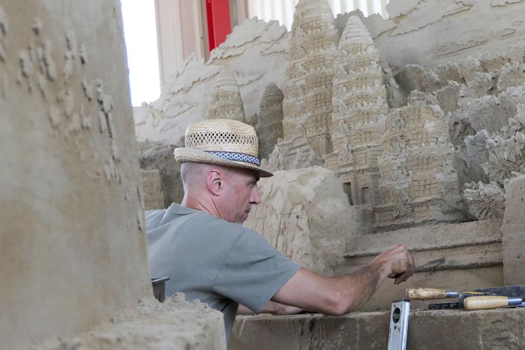 Sandskulpturenausstellung: Mann mit Strohhut formt filigrane Tortürme einer Tempelanlage aus verdichtetem Sand.