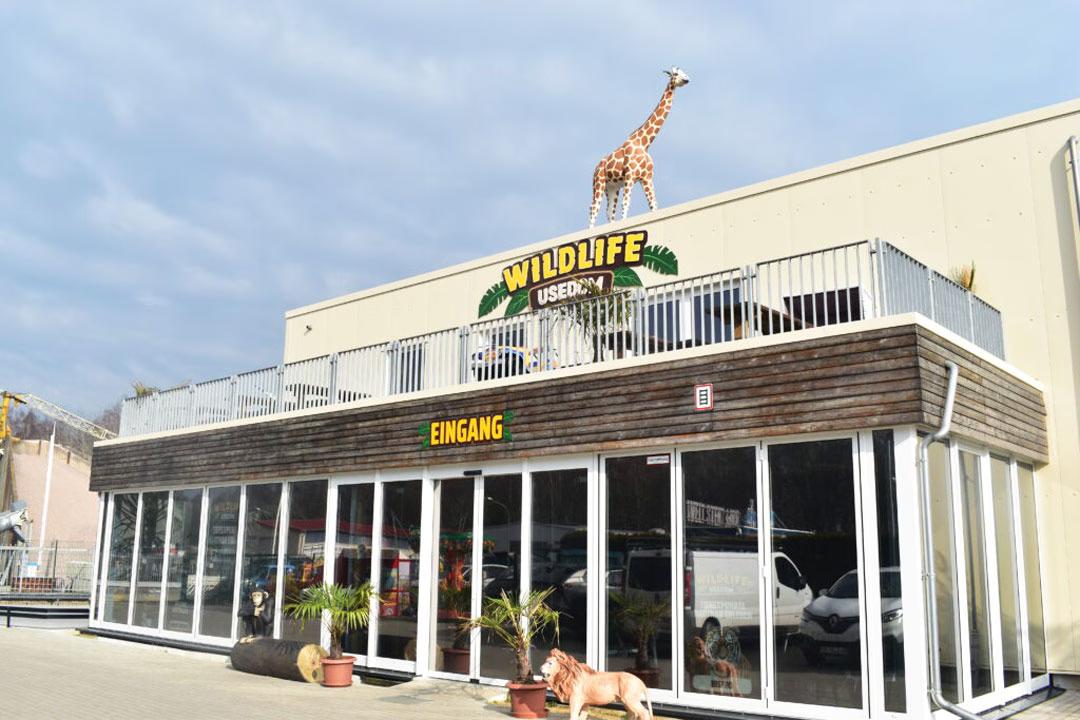 Außenansicht des Wildlife Usedom Gebäudes mit Eingangsbereich, großen Glasfronten und lebensgroßer Giraffe auf dem Dach.