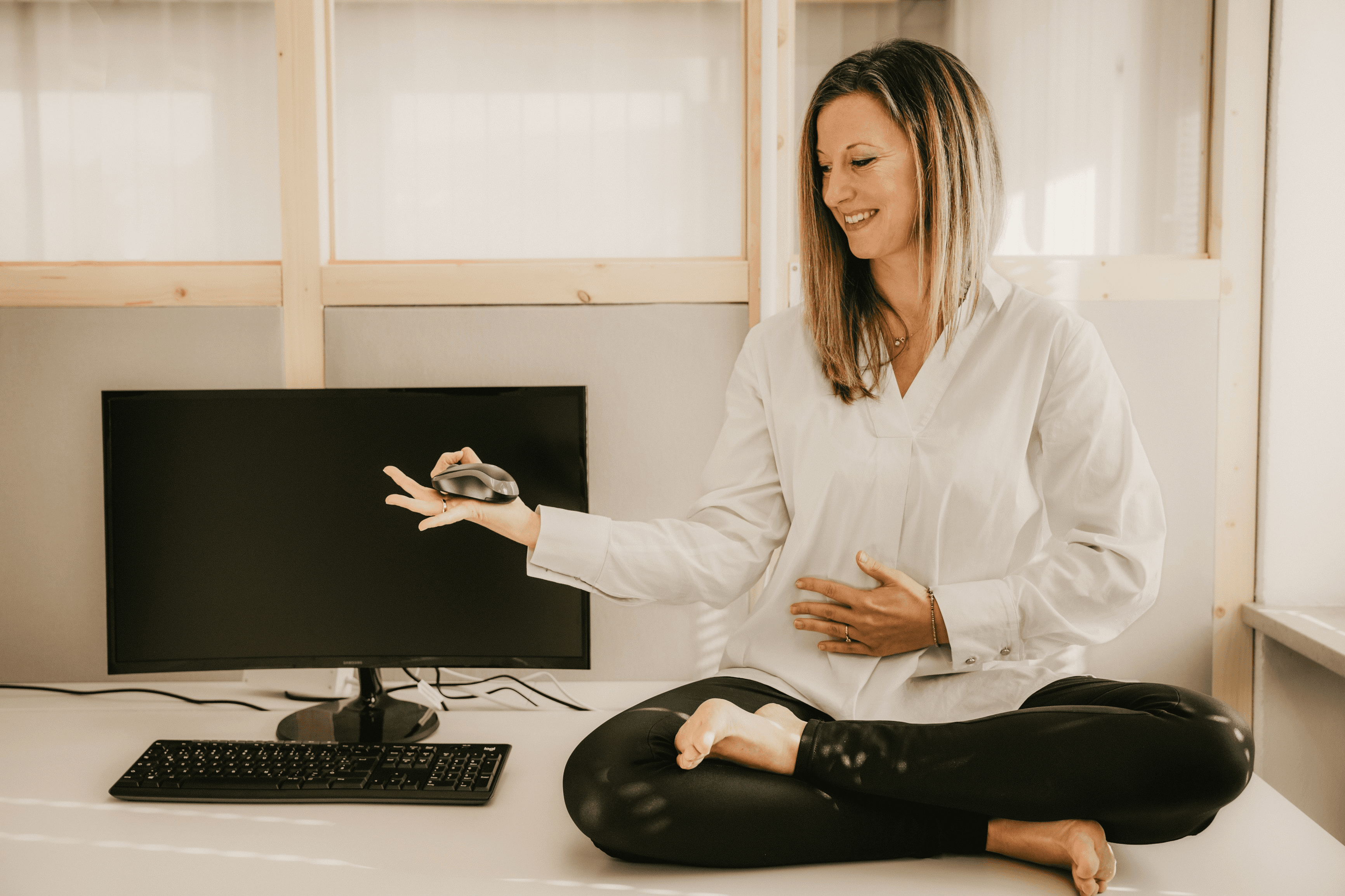 Die Trainerin Dr. Martina Weifenbach sitzt im Lotos-Sitz auf einem Schreibtisch und hält eine Computermaus in der Hand