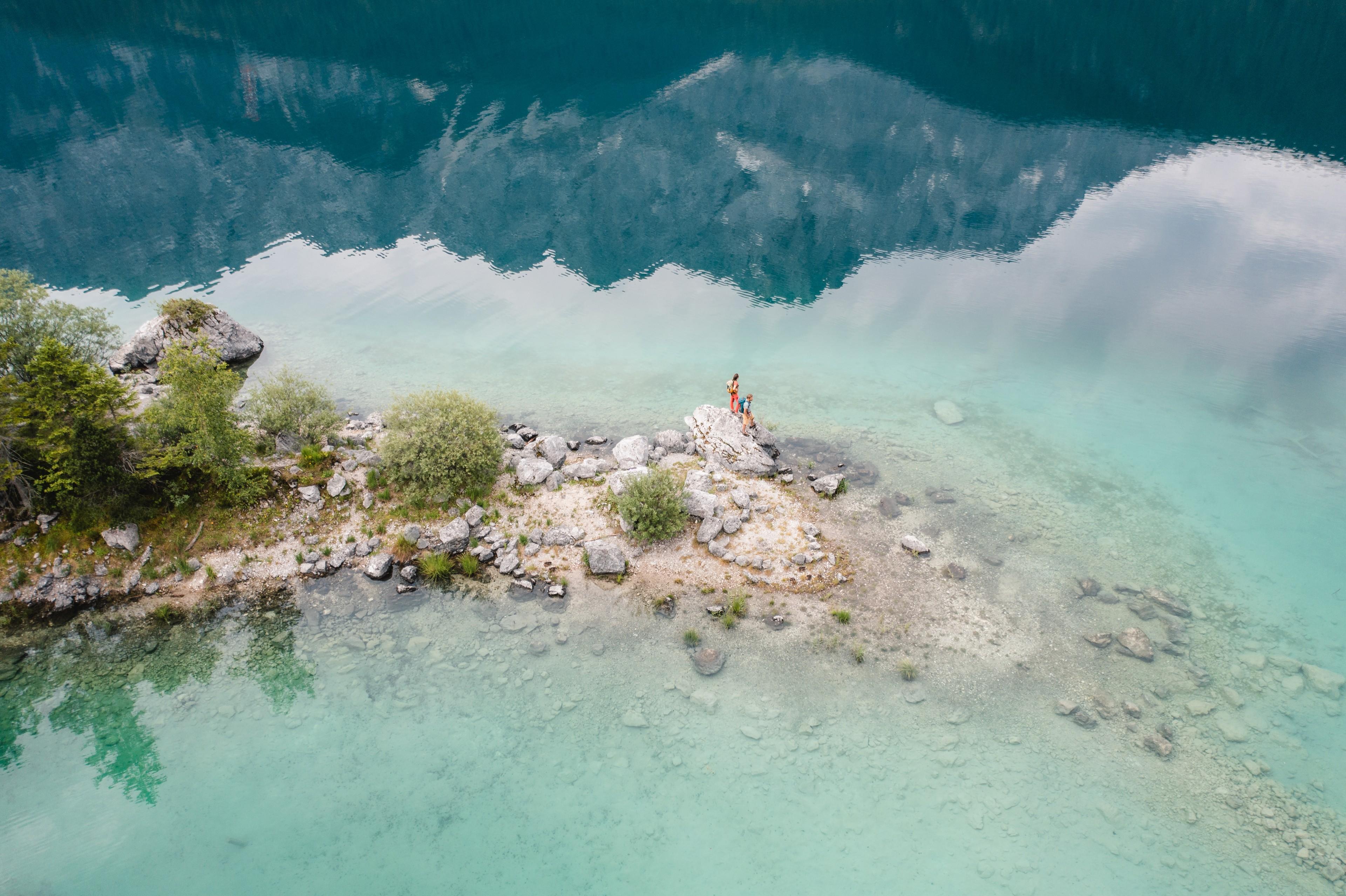 Zwei Urlauber:innen sitzen am Eibsee, die Alpen spiegeln sich im Wasser