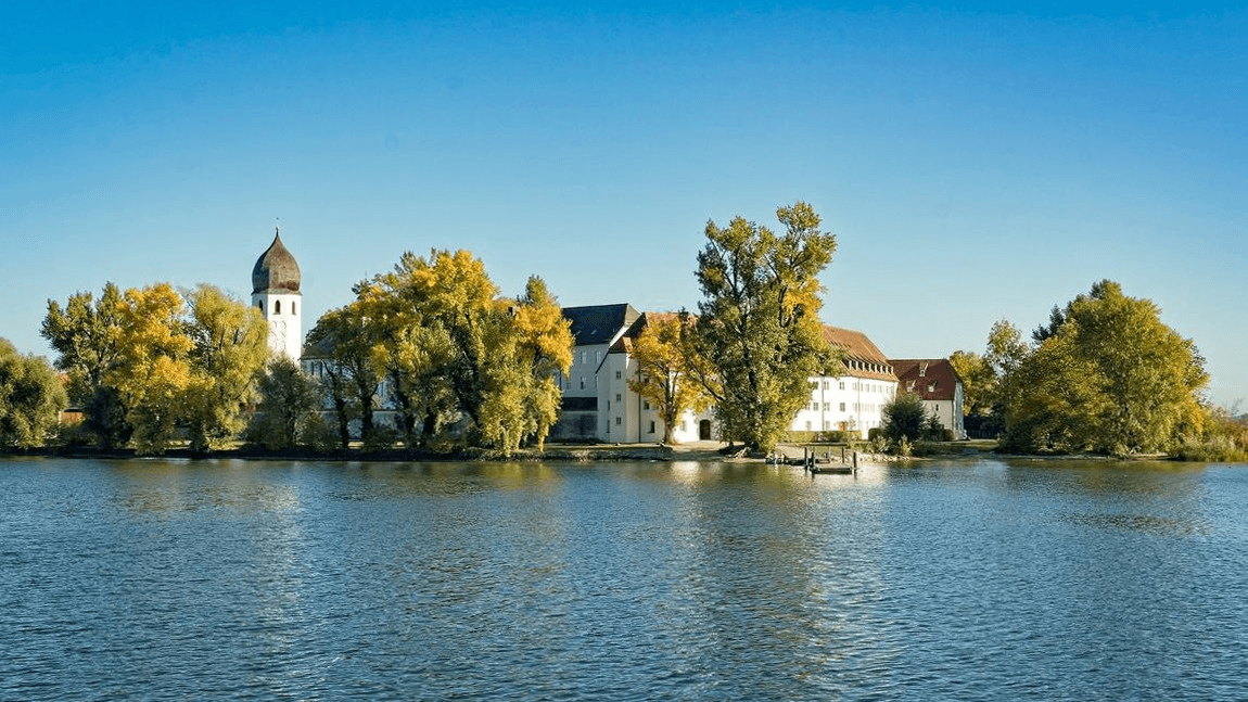 Die weißen Klostermauern von Frauenwörth im Chiemsee hinter Bäumen vom Wasser aus gesehen