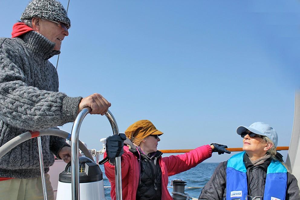 Der Steuermann eines Schiffes unterhält sich mit zwei Teilnehmenden eines Segeltörns