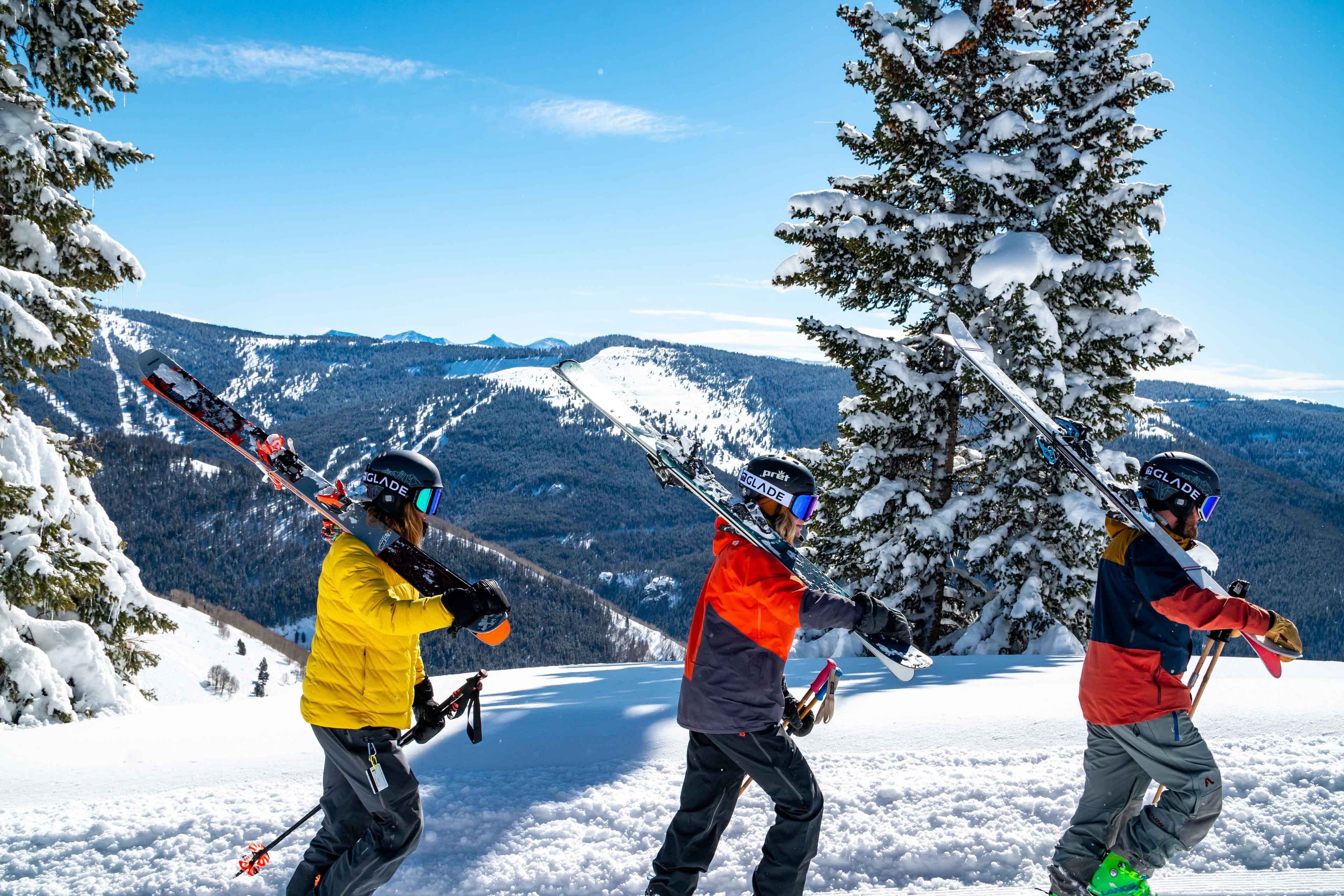 Drei Skifahrer:innen stapfen mit geschulterten Ski durch den Schnee