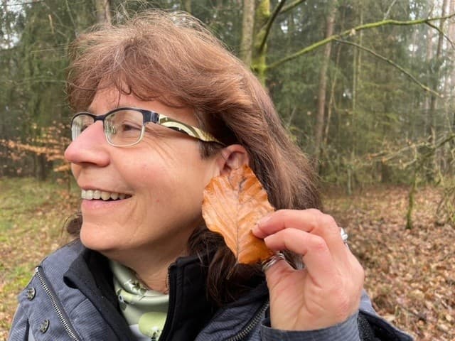 Eine Frau hält bei einem Waldbaden-Retreat ein Blatt an ihr Ohr und lauscht dem Knistern