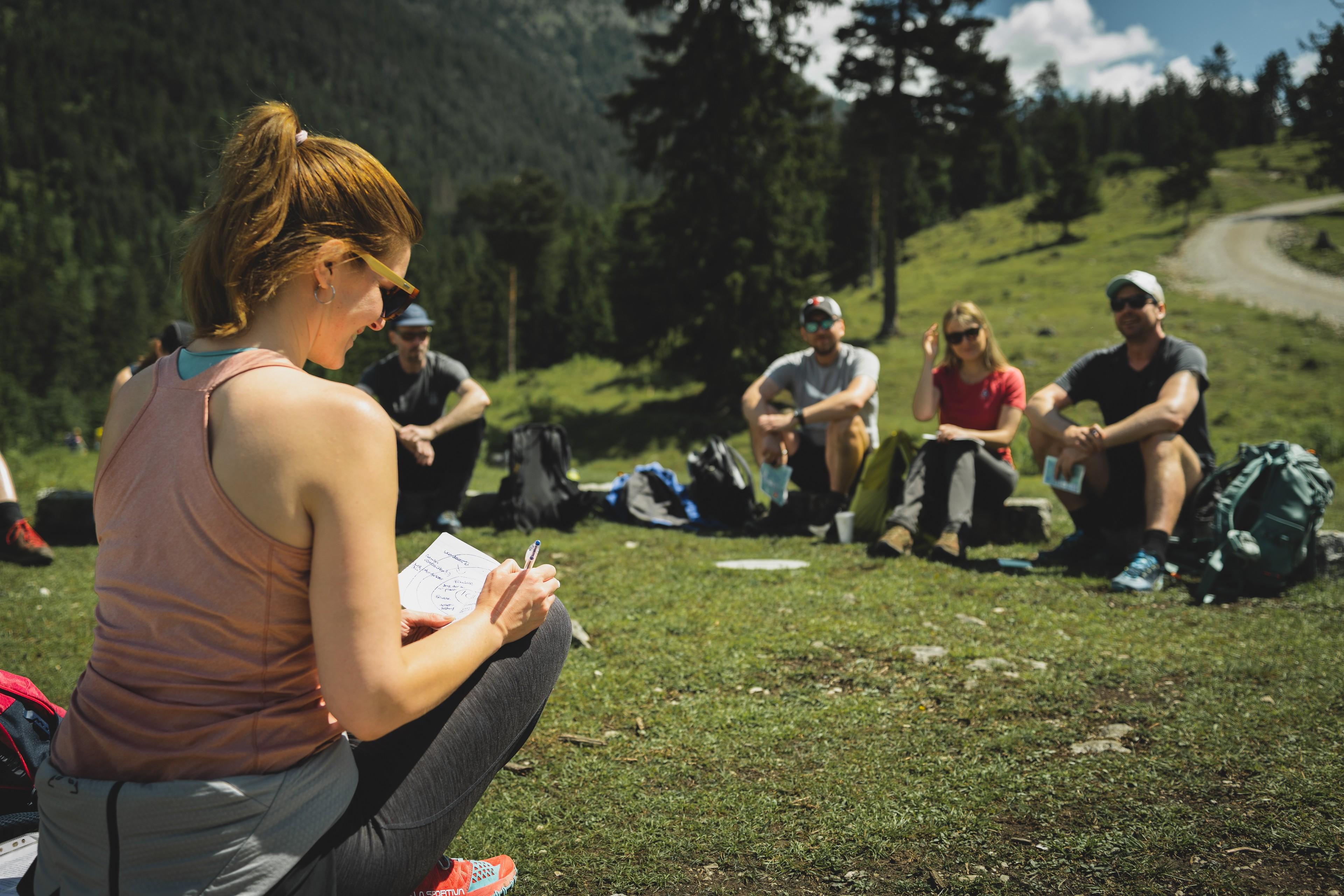 Ein weiblicher Coach sitzt vor einer Gruppe auf einer Wiese in den Alpen