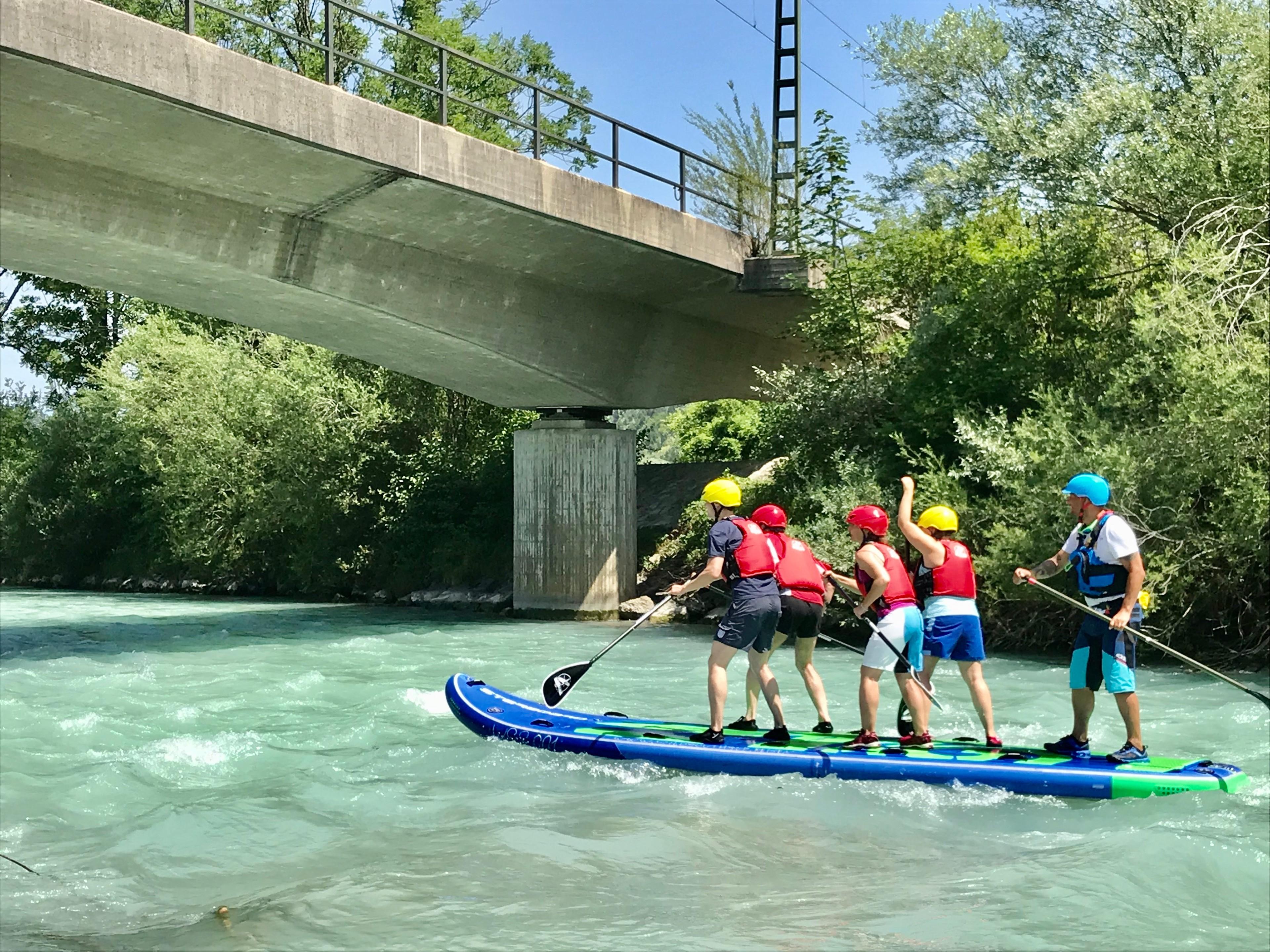 Eine Gruppe Menschen steht auf einem Maxi-SUP-Board und paddelt auf einem Fluss unter einer Brücke hindurch