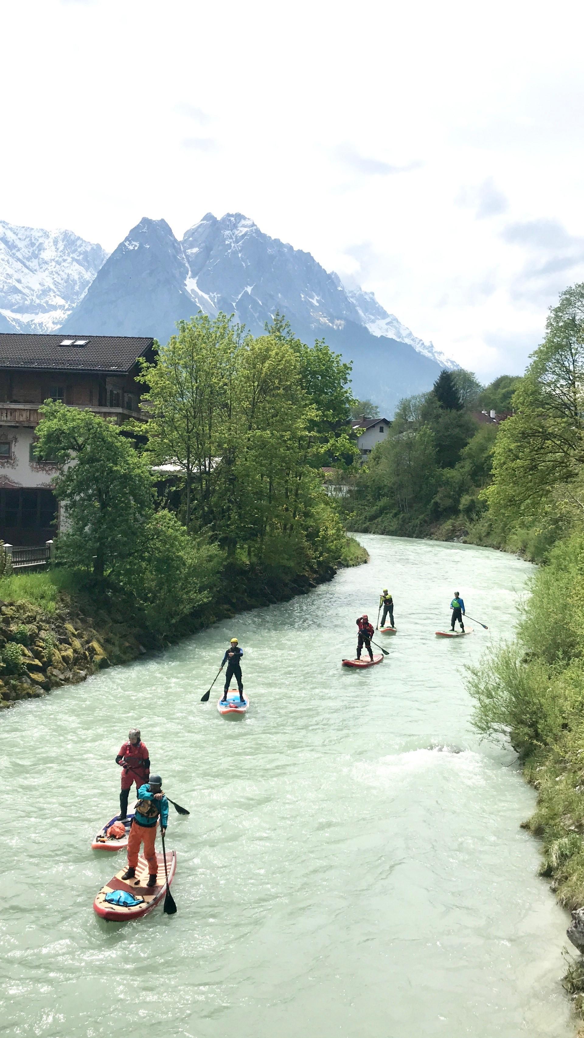 Eine Gruppe von Menschen auf SUP-Boards paddelt auf der Loisach mit den Alpen im Hintergrund