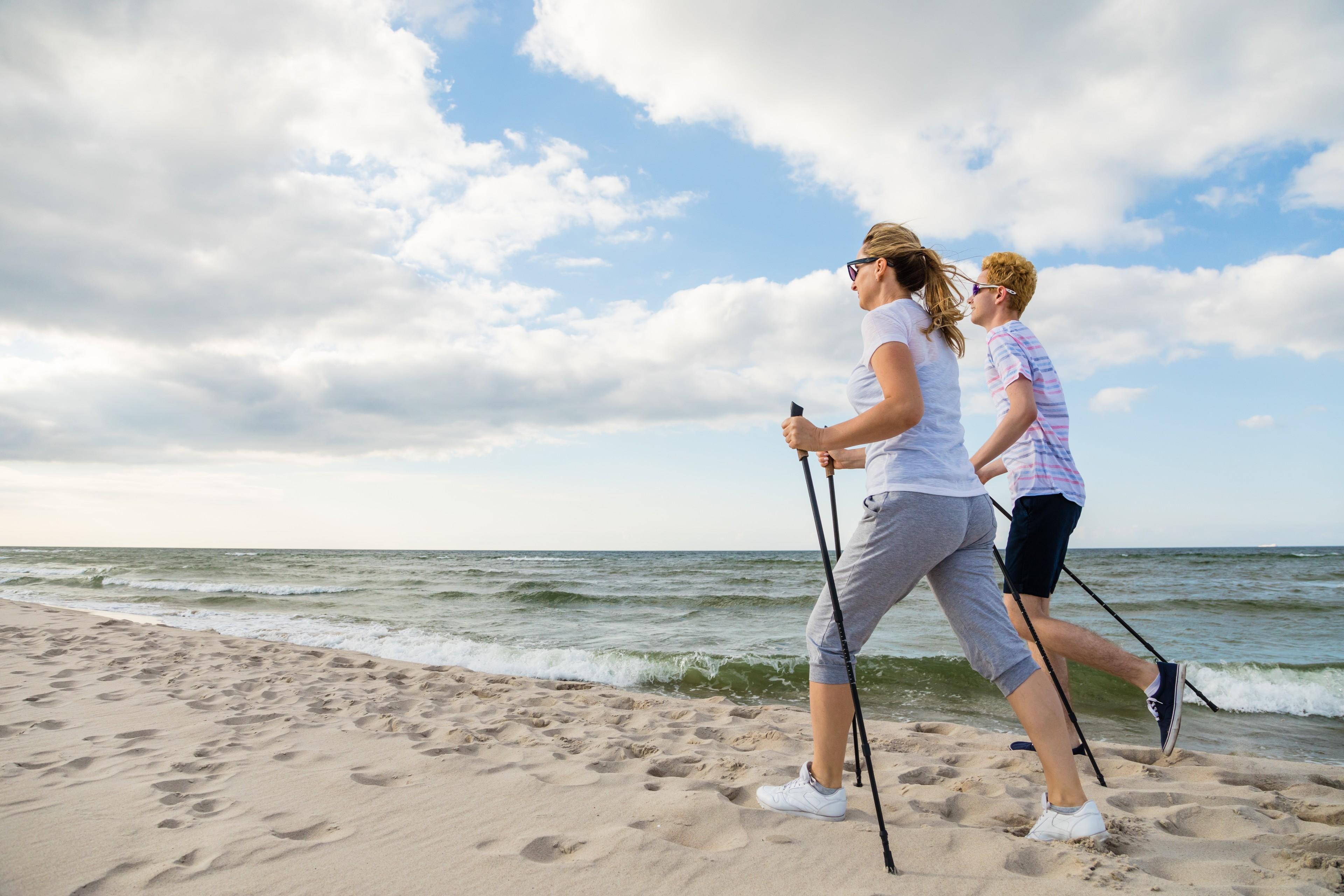 2 Personen machen in Sportkleidung und mit Stöcken Nordic Walking am Strand